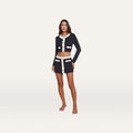 A woman stands barefoot against a plain white background, wearing the Adaline Cardigan Cover-Up—a matching navy blue cropped jacket and mini skirt set with white trim and gold buttons—perfect as a stylish swimsuit cover-up from beach to boat.