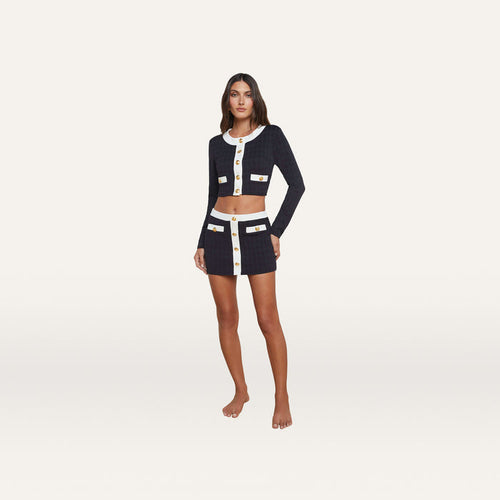 A woman stands barefoot against a plain white background, wearing the Adaline Cardigan Cover-Up—a matching navy blue cropped jacket and mini skirt set with white trim and gold buttons—perfect as a stylish swimsuit cover-up from beach to boat.