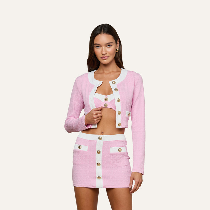 Wearing the Adaline Cardigan Cover-Up in light pink with white trim and gold buttons, paired with a matching pink textured skirt and crop top, she creates a stylish and versatile look that’s perfect for beach-to-boat outings against a plain white background.