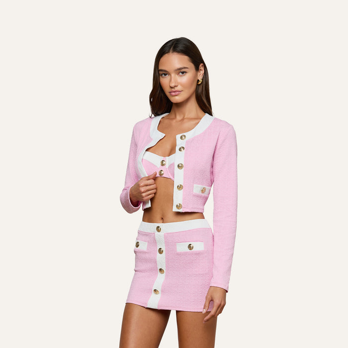 Wearing the Adaline Cardigan Cover-Up in pink and white, a woman poses against a plain white background. She styles the buttoned crop jacket with its matching bralette and high-waisted skirt for a chic swimsuit cover-up look.