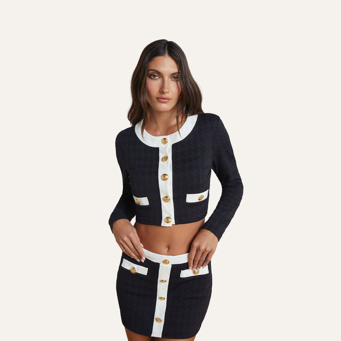 Wearing the Adaline Cardigan Cover-Up styled as a chic beach-to-boat look, a woman poses against a plain light background in this black cropped jacket and matching skirt set, featuring white trim and gold buttons.