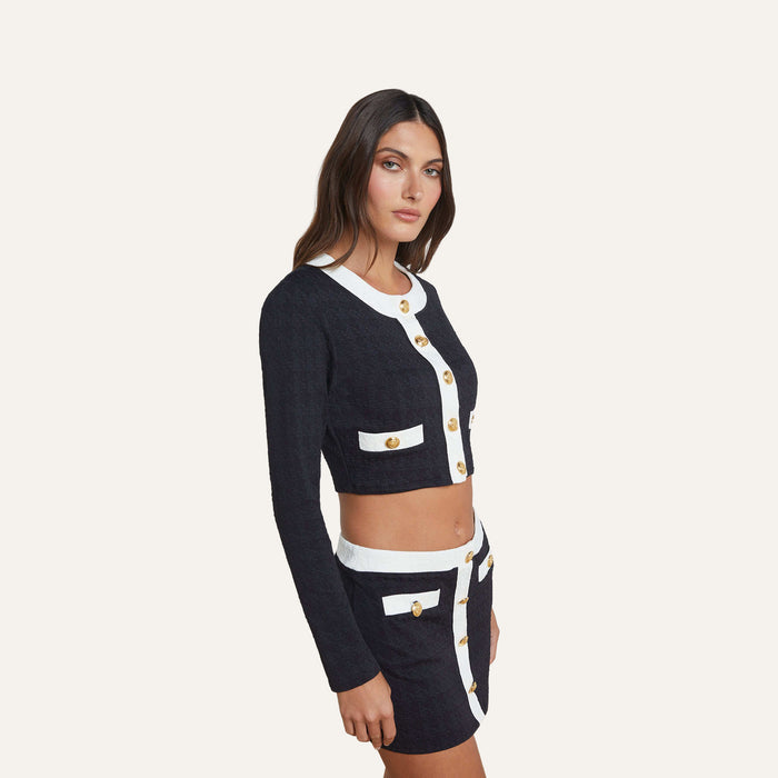 Wearing the Adaline Cardigan Cover-Up—a black and white long-sleeve cropped jacket with gold buttons that’s perfect as a chic swimsuit cover-up—paired with a high-waisted mini skirt, she poses against a plain light background.