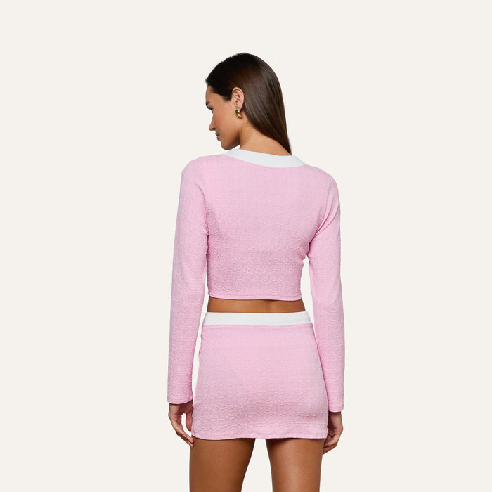 A woman is standing with her back to the camera, wearing the Adaline Cardigan Cover-Up—a light pink long-sleeve crop top and matching mini skirt with white trim—styled as a chic swimsuit cover-up against a plain light background.