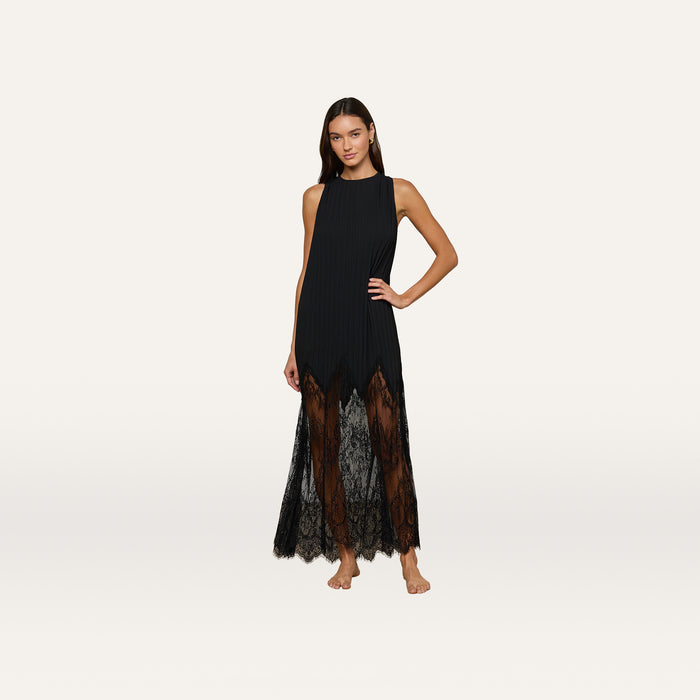 A woman stands barefoot against a plain light background, posing with one hand on her hip and wearing the Adalynn Cover-Up Dress, a sleeveless black lace style featuring sheer lace details at the hem.