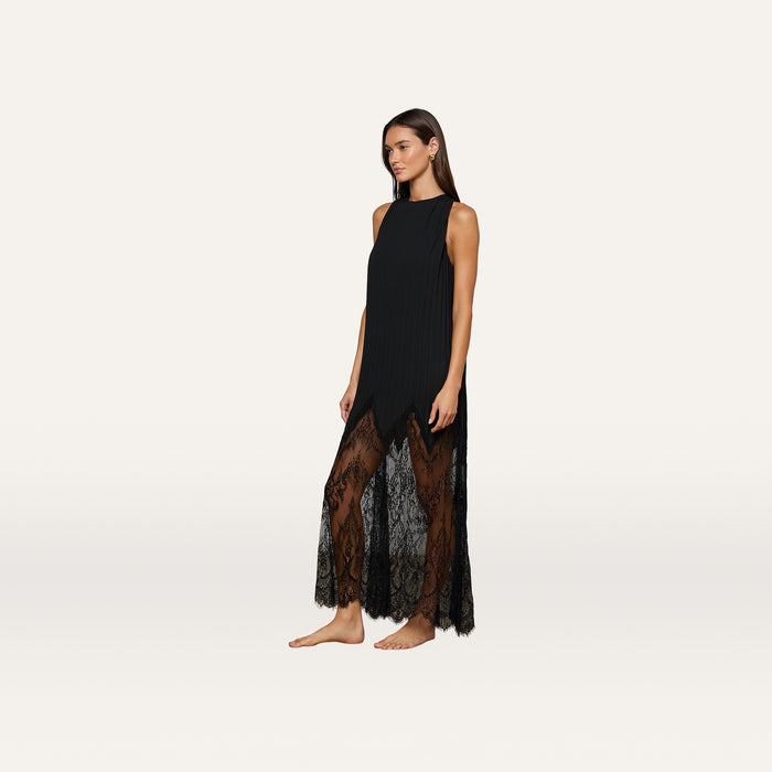 A woman stands barefoot against a plain, light background wearing the Adalynn Cover-Up Dress—an ankle-length, sleeveless black lace dress featuring sheer detailing at the hem.