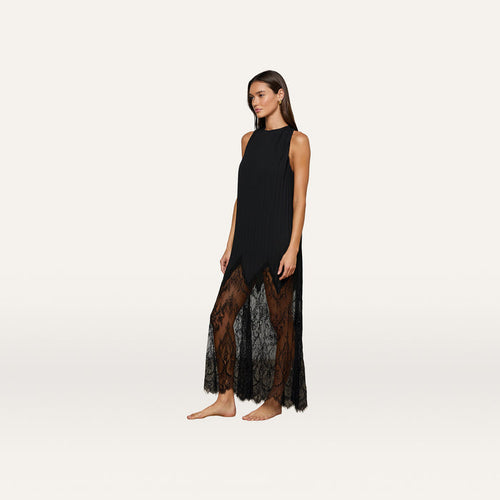 A woman stands barefoot against a plain, light background wearing the Adalynn Cover-Up Dress—an ankle-length, sleeveless black lace dress featuring sheer detailing at the hem.