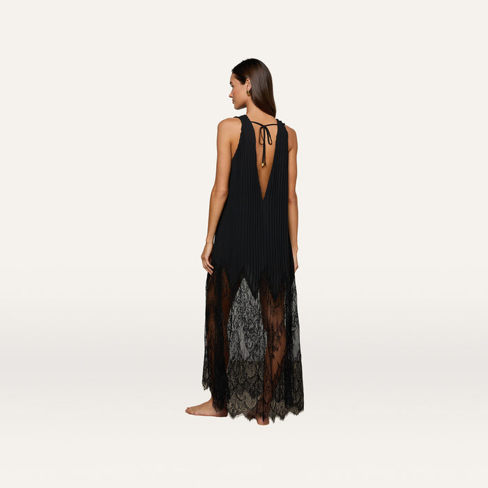 A woman stands barefoot and facing away on a plain light background, wearing the Adalynn Cover-Up Dress—a sleeveless black lace dress featuring a deep V-back, pleated upper section, chiffon lower skirt, and a tie detail at the neck.