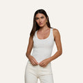 Woman with long brown hair wearing the Adira Scoopneck Tank and off-white pants, standing against a plain light background, looking at the camera with a neutral expression.