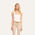 A woman with long blonde hair wears the Adira Scoopneck Tank in white, paired with beige pants, a tan belt, and gold earrings, standing against a plain light background.