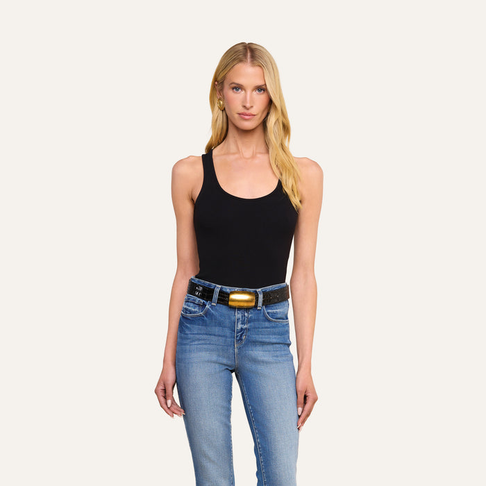 A woman with long blonde hair wearing the slim fit Adira Scoopneck Tank in black and blue jeans with a black belt featuring a large oval buckle stands against a plain light background.
