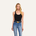 A woman with long blonde hair wearing the slim fit Adira Scoopneck Tank in black and blue jeans with a black belt featuring a large oval buckle stands against a plain light background.
