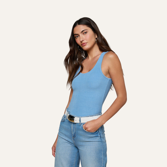Woman with long brown hair wearing the Adira Scoopneck Tank in blue, paired with light blue jeans and a white belt, standing against a plain light background with one hand in her pocket.
