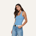 Woman with long brown hair wearing the Adira Scoopneck Tank in blue, paired with light blue jeans and a white belt, standing against a plain light background with one hand in her pocket.