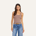 A woman stands facing forward, wearing the Adira Scoopneck Tank in taupe, paired with blue jeans and a tan belt. She has long, wavy brown hair and a neutral expression against a plain, light background.