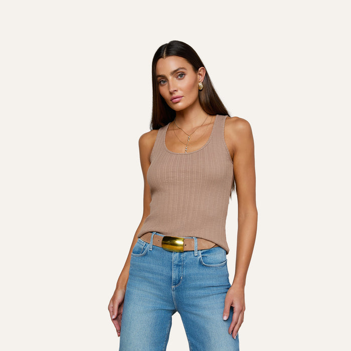 A woman with long brown hair wears the Adira Scoopneck Tank—a fitted, sleeveless beige ribbed top crafted from organic cotton—paired with blue jeans and a belt. She stands against a plain light background, posing with one hand in her pocket and looking toward the camera.