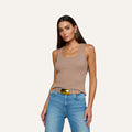 A woman with long brown hair wears the Adira Scoopneck Tank—a fitted, sleeveless beige ribbed top crafted from organic cotton—paired with blue jeans and a belt. She stands against a plain light background, posing with one hand in her pocket and looking toward the camera.