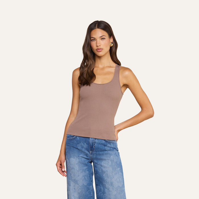 A woman with long brown hair wears the Adira Scoopneck Tank— a sleeveless taupe essential top—paired with blue wide-leg jeans, standing against a plain light background with one hand on her hip.