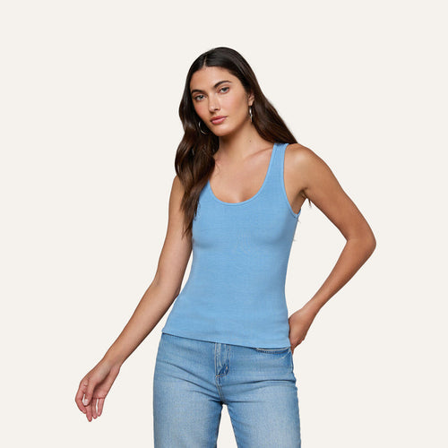 A woman with long brown hair wears the Adira Scoopneck Tank in light blue and blue jeans, standing against a plain light background with one arm relaxed and the other bent at the elbow.