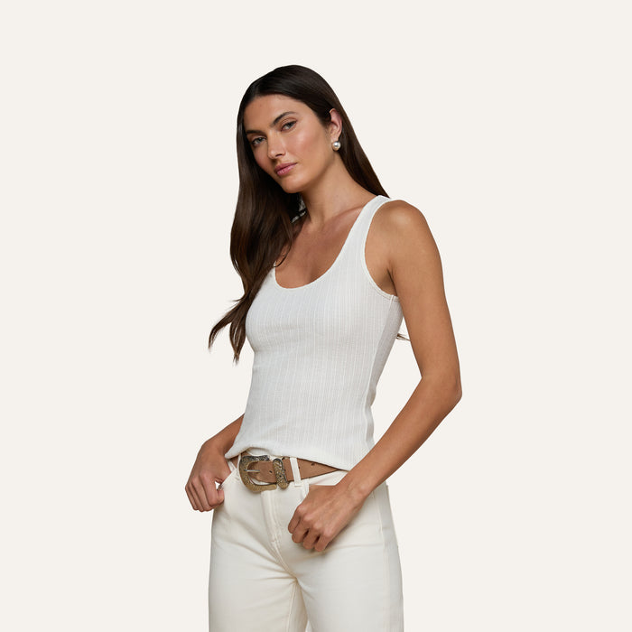 A woman with long dark hair wears the Adira Scoopneck Tank in white, paired with white pants and a brown belt, standing against a plain light background.