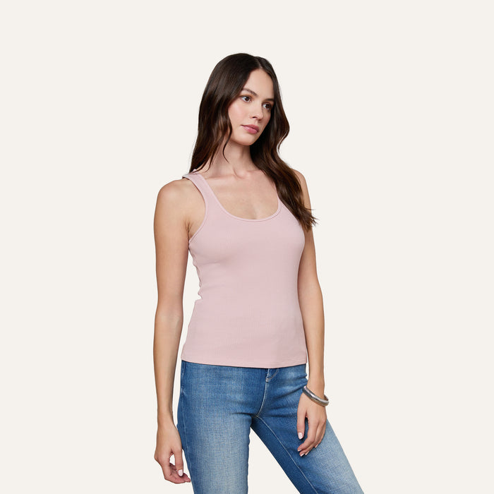 A woman with long brown hair wears the Adira Scoopneck Tank in light pink and blue jeans, standing against a plain light background with a neutral expression.