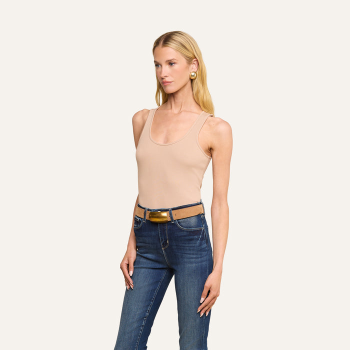 A woman with long blonde hair, wearing the Adira Scoopneck Tank in beige ribbed stretch-cotton, blue jeans, a brown belt, and large gold earrings stands against a plain light background.
