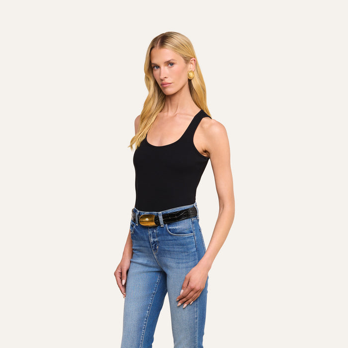 A woman with long blonde hair wears the Adira Scoopneck Tank in black, paired with blue jeans, a black belt with a gold buckle, and gold earrings, standing against a plain light background.