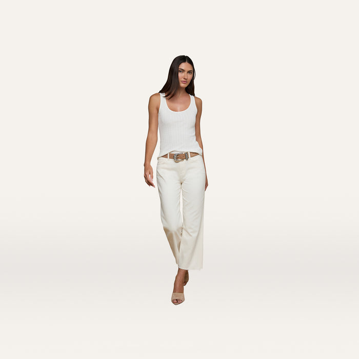 A woman stands against a plain light background, wearing the Adira Scoopneck Tank in white, wide-leg white pants, a brown belt, and nude heels.