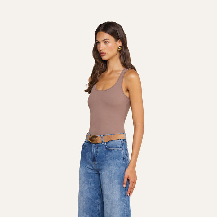 A woman with long brown hair wears the Adira Scoopneck Tank in taupe, paired with wide-leg blue jeans featuring a floral pattern and a brown belt with a gold buckle. She stands facing left against a plain background.