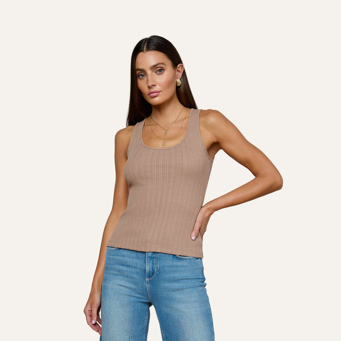 A woman wearing the Adira Scoopneck Tank—a fitted, ribbed beige organic cotton top—paired with blue jeans stands against a plain light background, one hand on her hip and a neutral expression.