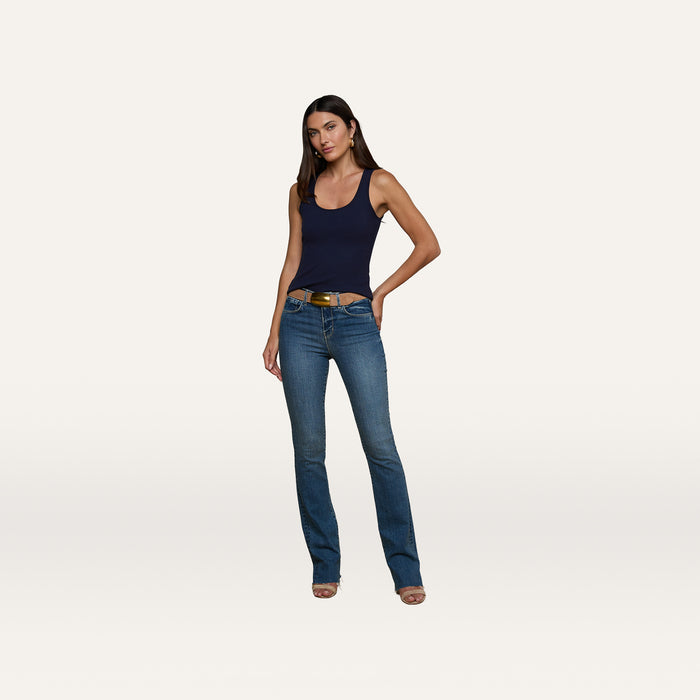 A woman stands against a plain white background, wearing the Adira Scoopneck Tank in essential navy stretch rib fabric, blue jeans with a brown belt, and beige heeled sandals. She has straight dark hair and her left hand is on her hip.