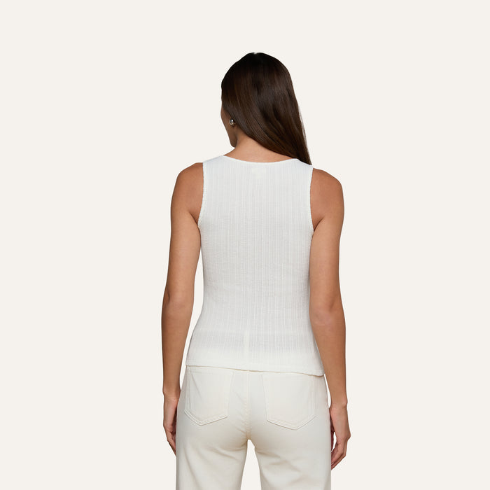 A woman stands facing away, wearing the Adira Scoopneck Tank—a sleeveless white cotton pointelle top—paired with white pants against a plain light background.