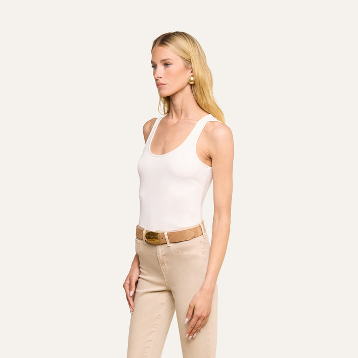 A woman stands against a plain background, wearing the Adira Scoopneck Tank in white, beige pants with a belt, and gold earrings. She has long blonde hair and a neutral expression, facing slightly to the left.