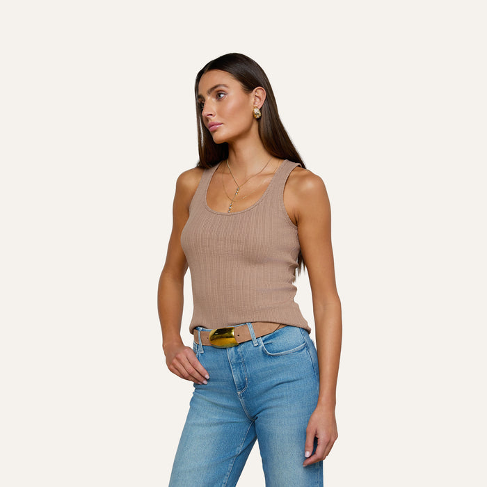 Wearing the Adira Scoopneck Tank—a fitted tan ribbed top—paired with blue jeans and a belt, a woman stands against a plain light background, gazing slightly to the side with one hand in her pocket.