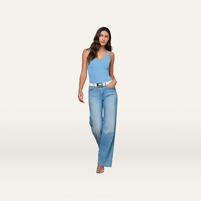 A woman stands against a plain background, wearing the Adira Scoopneck Tank in light blue, wide-leg blue jeans, a white belt, and heeled sandals. She has long, wavy hair and a neutral expression.