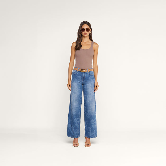 Wearing sunglasses, wide-leg blue jeans with a belt, and heeled sandals, a woman stands against a plain white background showcasing the Adira Scoopneck Tank in sleeveless beige.
