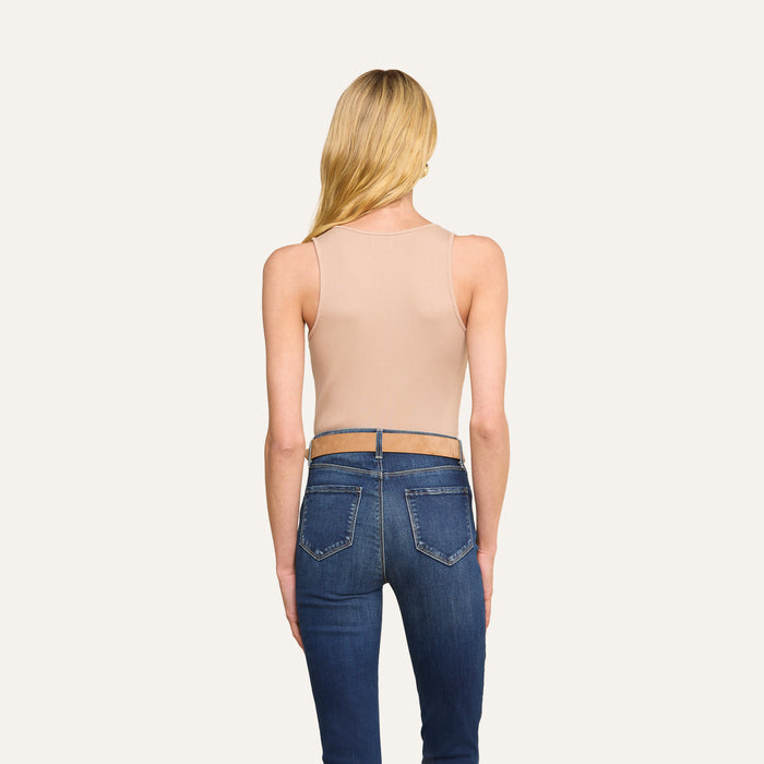 A woman with long blonde hair, wearing the Adira Scoopneck Tank in beige and blue jeans, stands facing away from the camera against a plain light background.