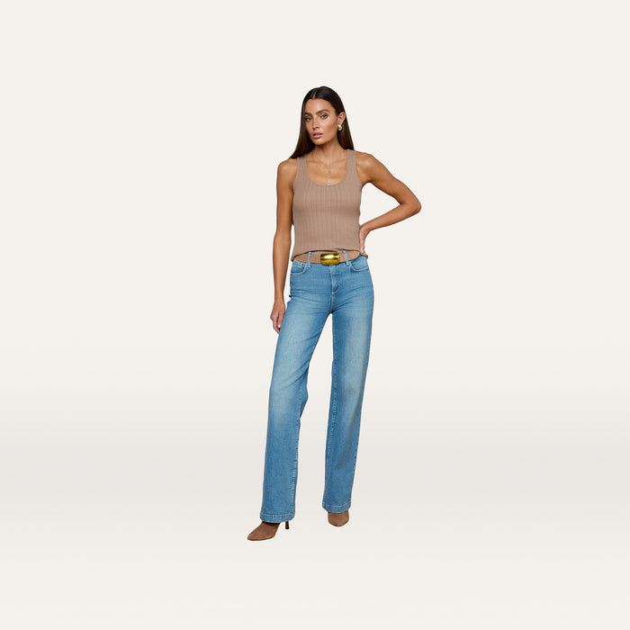 A woman models the Adira Scoopneck Tank— a fitted, sleeveless beige top with a scoop neckline— paired with blue jeans and brown heels, standing with one hand on her hip against a plain white background.