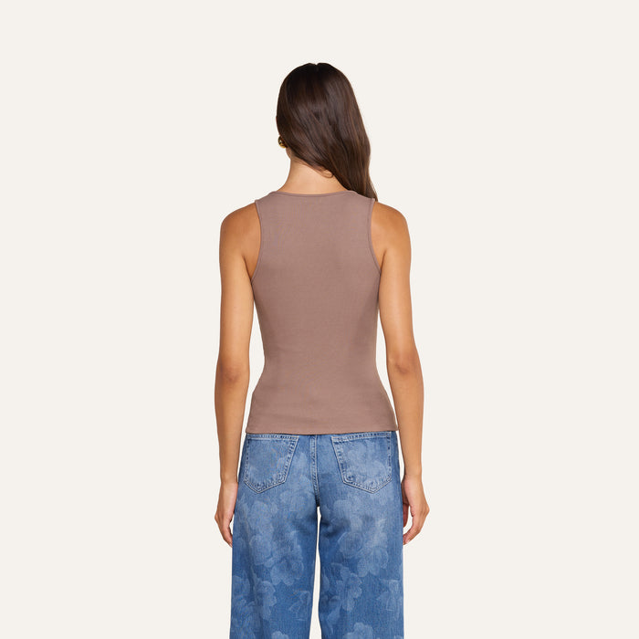 A woman stands facing away, wearing the Adira Scoopneck Tank in brown and blue jeans with a floral pattern on the back pockets, against a plain light background.