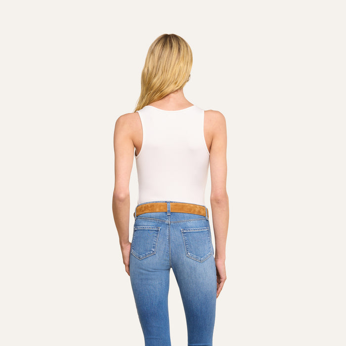 A woman with long blonde hair, wearing the Adira Scoopneck Tank in white and blue jeans with a brown belt, stands facing away from the camera against a plain light background.