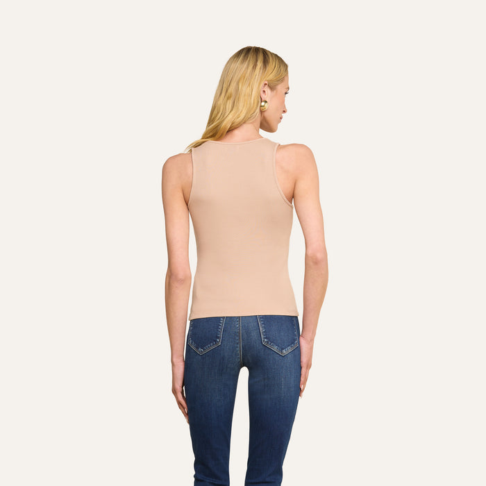 A person with long blonde hair, wearing the Adira Scoopneck Tank in sleeveless beige and blue jeans, stands facing away from the camera against a plain light background.
