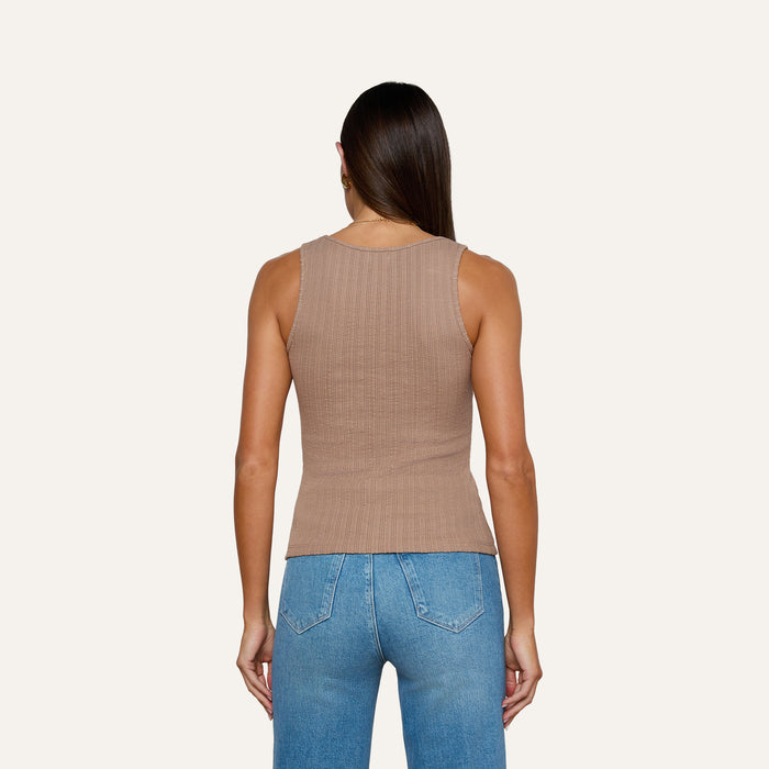 A woman with long brown hair, wearing the Adira Scoopneck Tank—a fitted, sleeveless beige top made from organic cotton—and blue jeans stands with her back to the camera against a plain light background.