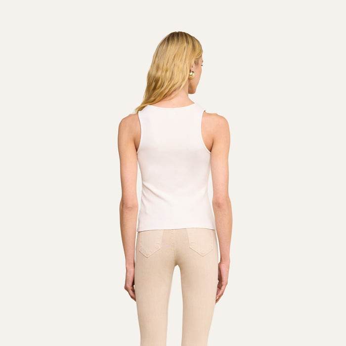 A woman with long blonde hair, wearing the Adira Scoopneck Tank in white and light beige pants, stands facing away from the camera against a plain light background.