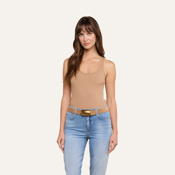 A woman with long brown hair wears the Adira Tank in tan, styled with light blue jeans and a belt with a gold buckle, while standing against a plain light background.