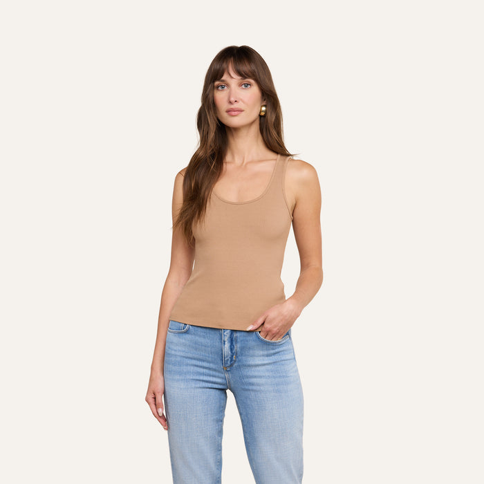 A woman with long brown hair wears the Adira Tank in tan, paired with light blue jeans, standing against a plain light background with one hand in her pocket.