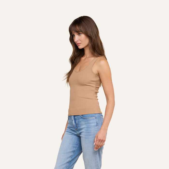 A woman with long brown hair wearing the Adira Tank in tan and light blue jeans stands against a plain light background, facing left with a neutral expression.