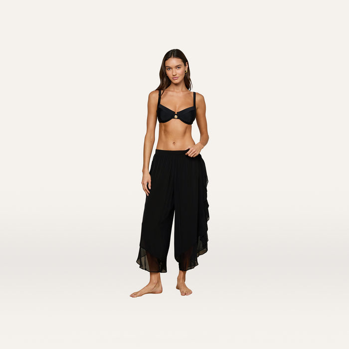 A woman stands barefoot against a plain light background, wearing a black bikini top and the Adonis Cover-Up Pant—loose, flowy pants made from sheer chiffon.