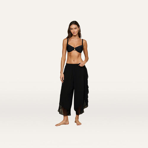 A woman stands barefoot against a plain light background, wearing a black bikini top and the Adonis Cover-Up Pant—loose, flowy pants made from sheer chiffon.