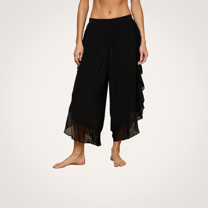 A person is wearing the Adonis Cover-Up Pant—loose-fitting, black, semi-sheer chiffon ruffled capri pants that double as a stylish cover-up—standing barefoot against a plain, light background.