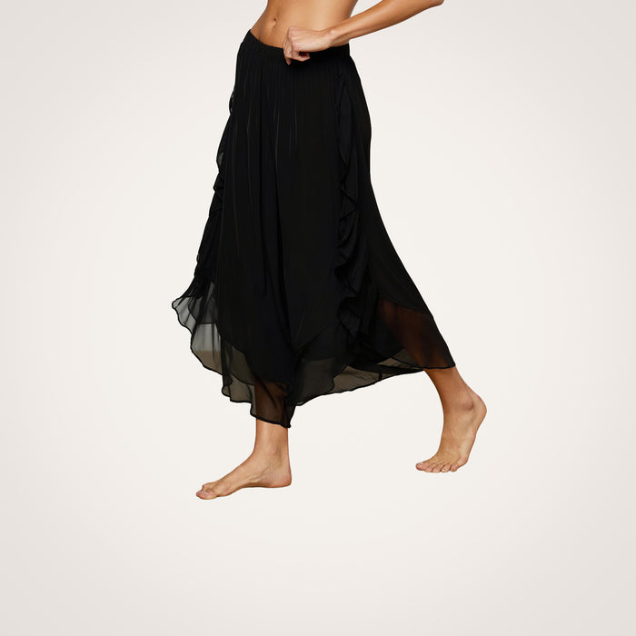 A person wears the Adonis Cover-Up Pant, a flowing black sheer chiffon midi skirt with an elastic waistband, while walking barefoot against a light background. Only the lower torso and legs are visible.