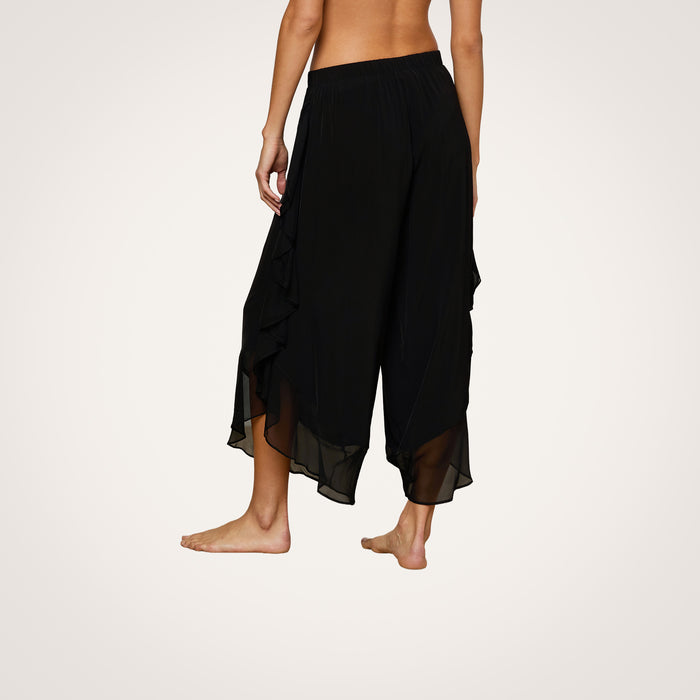 A person stands barefoot, showcasing the Adonis Cover-Up Pant—a pair of loose, black flowy pants with ruffled side details crafted from sheer chiffon. The image captures the wearer from the waist down against a plain light background, evoking an effortless poolside style.